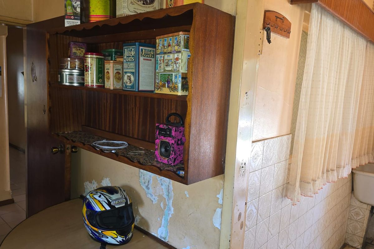 A dated kitchen area featuring built-in wooden shelving and ceramic wall tiling, currently showing significant wear and peeling paint on the walls.