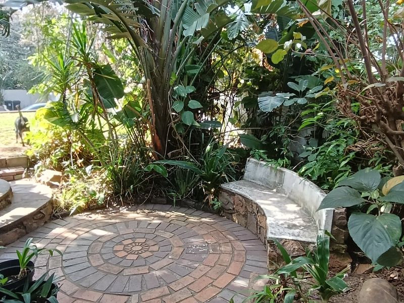A tranquil garden area featuring a circular stone-paved patio and a built-in stone bench surrounded by mature, lush landscaping.