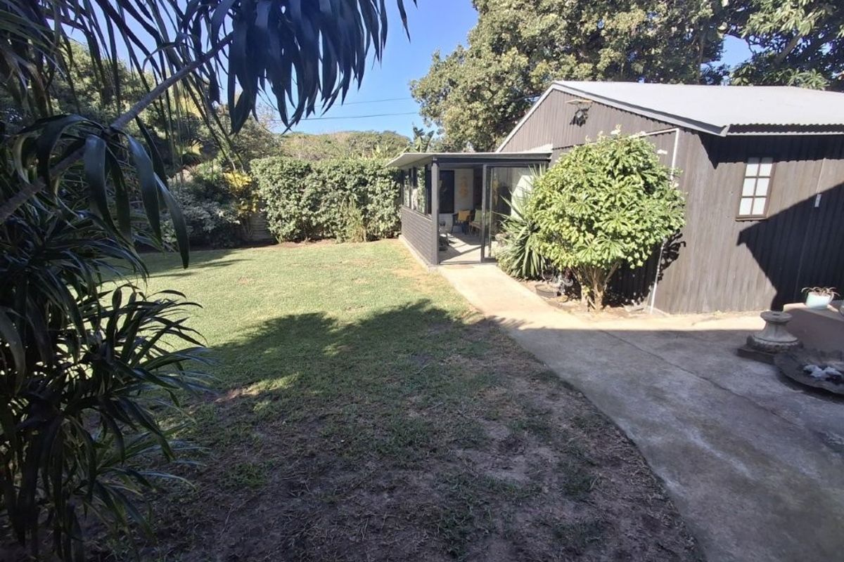 A rear garden view featuring a lawn, mature hedging, a concrete pathway, and a timber-clad structure with a covered patio area.