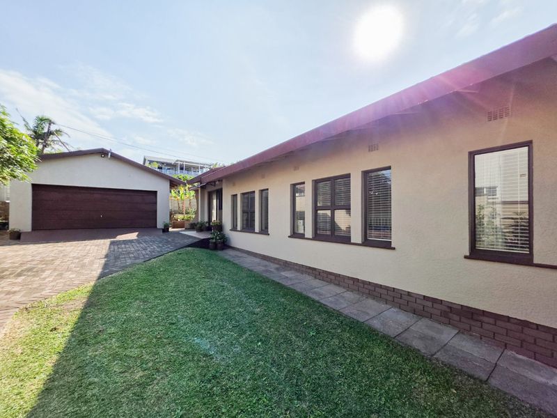 Rear view of the property featuring a paved driveway, detached garage, and a well-maintained lawn with a paved walkway along the building exterior.