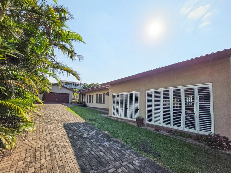 A well-maintained exterior view of the property featuring a paved driveway, landscaped garden, and white plantation shutters on the building facade.