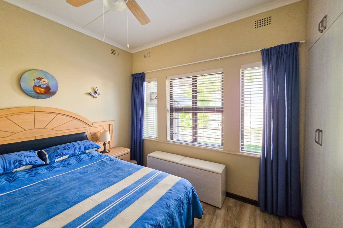 A well-lit bedroom featuring laminate flooring, built-in cupboards, a ceiling fan, and large windows with venetian blinds.