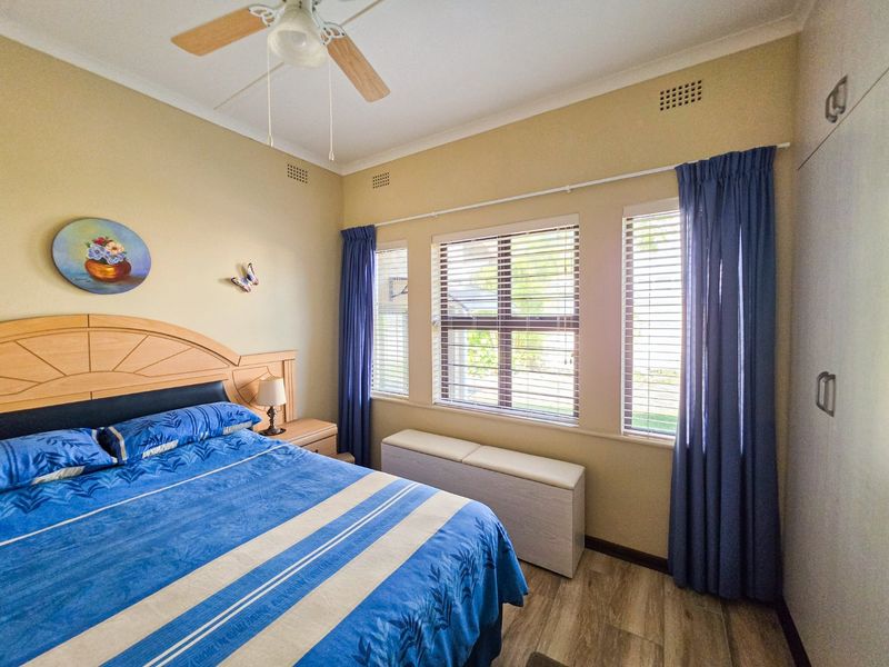 A well-lit bedroom featuring laminate flooring, built-in cupboards, a ceiling fan, and large windows with venetian blinds.