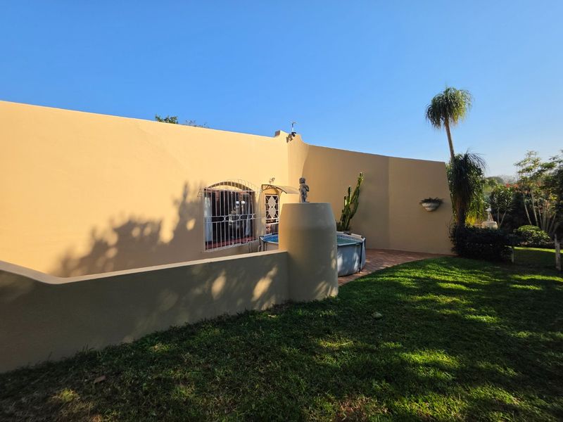 A view of the rear exterior of the house featuring a curved boundary wall, paved patio area, and well-maintained lawn.