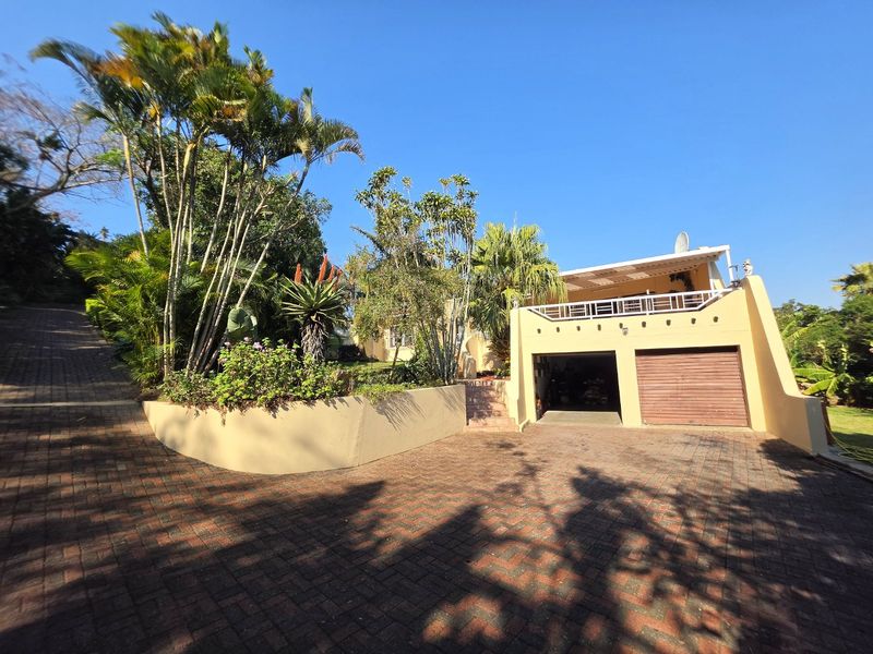 Front exterior view of the property featuring a paved driveway, double garage with metal doors, and an elevated balcony area with mature landscaping.