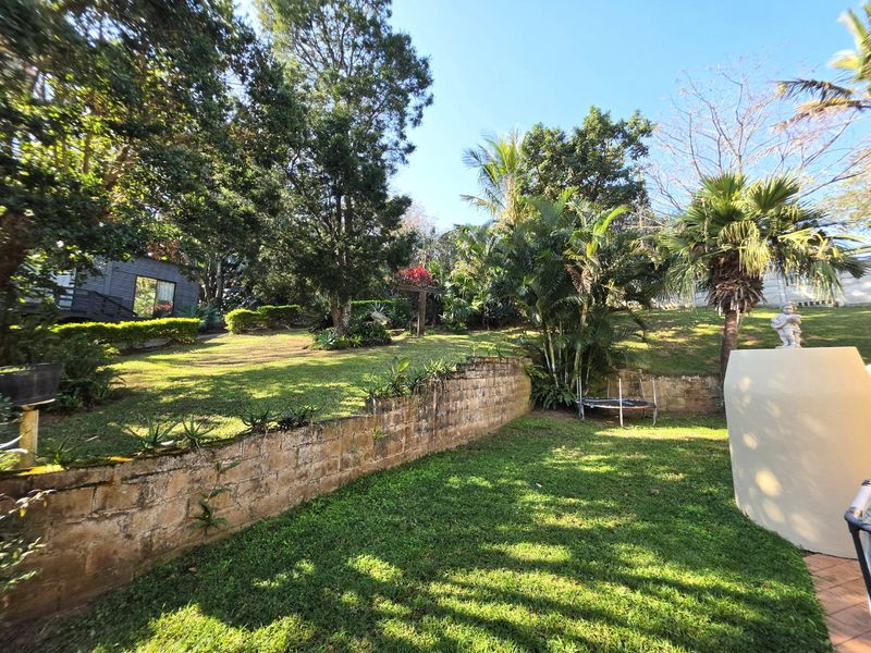 A spacious, multi-level garden featuring mature trees, established landscaping, and a stone retaining wall, providing a private outdoor setting.