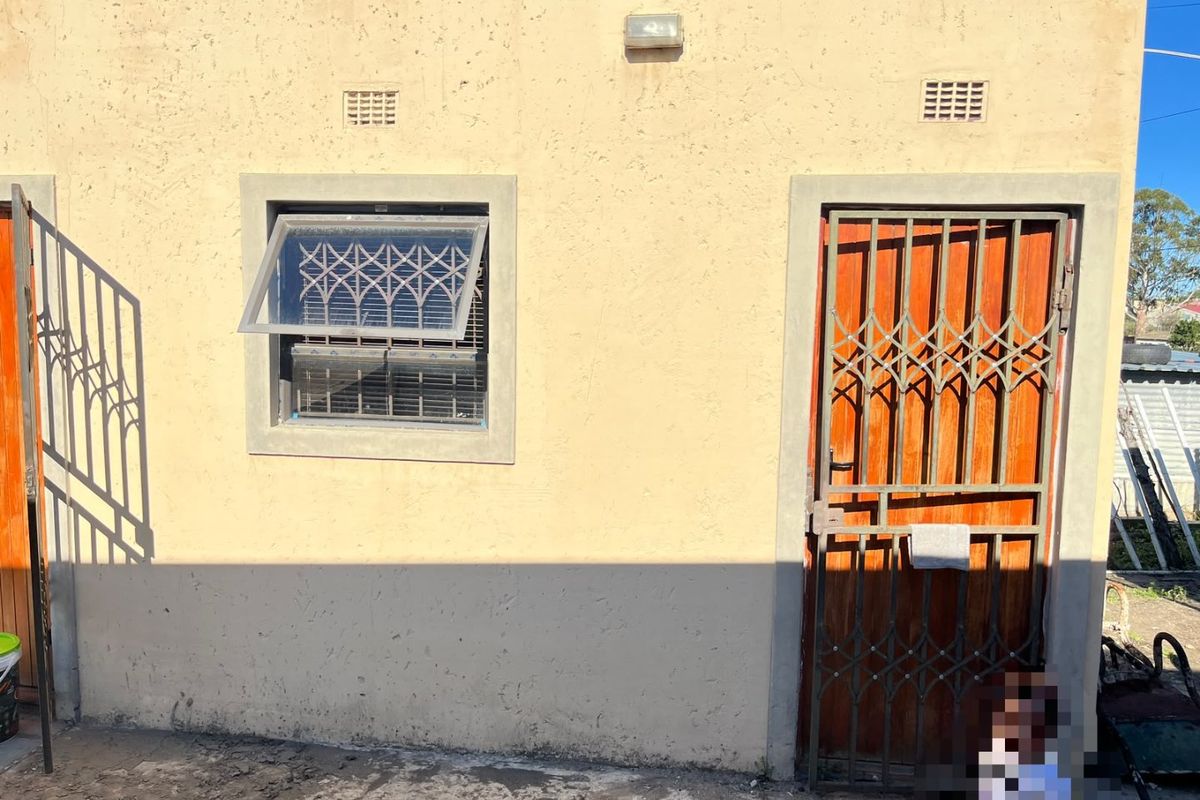 Rear exterior view of the property featuring a wooden door with a security gate, a top-hung window with security bars, and exterior wall lighting.