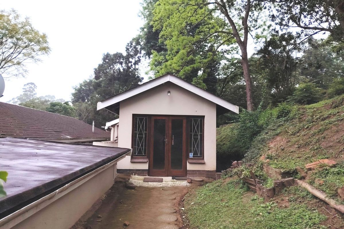 Rear view of a detached structure featuring a gabled roof, timber-framed glass doors, and leaded windows, set within a natural, sloped garden environment.