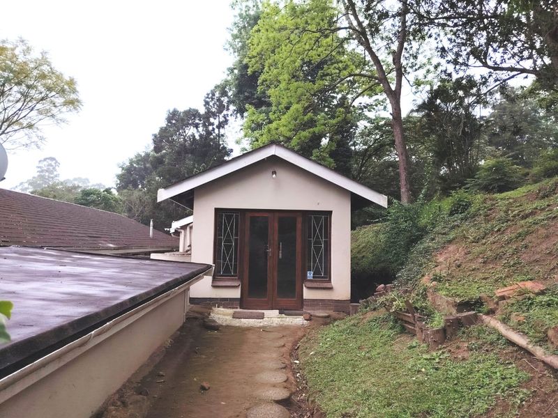 Rear view of a detached structure featuring a gabled roof, timber-framed glass doors, and leaded windows, set within a natural, sloped garden environment.