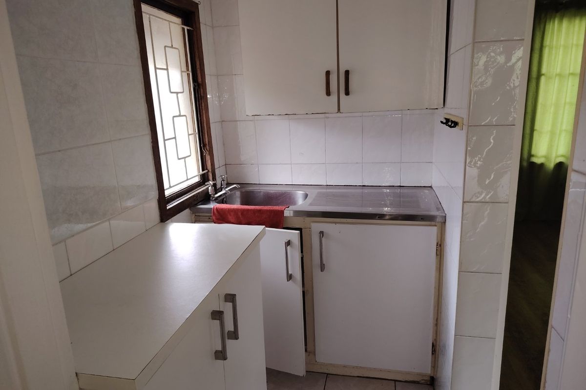 A compact kitchen featuring white built-in cabinetry, a stainless steel sink, and tiled wall finishes, requiring some maintenance and cosmetic updates.