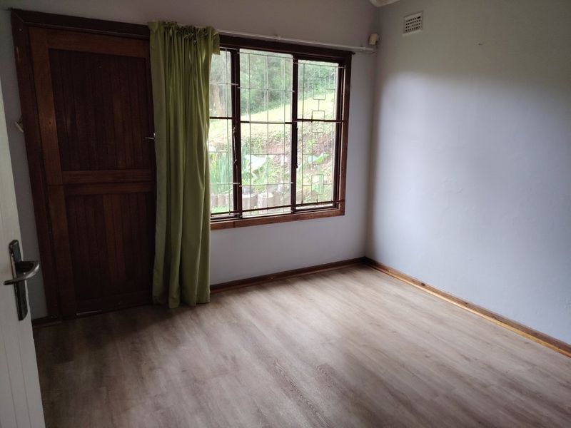 An empty bedroom featuring laminate flooring, a timber door, and a window with security bars, offering a view of the garden area.