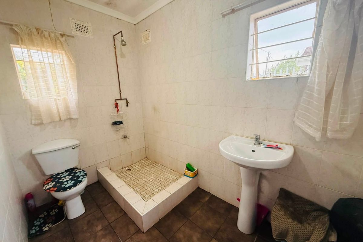 A functional bathroom featuring a pedestal basin, raised shower base with exposed plumbing, and tiled finishes, showing signs of wear and ceiling staining.