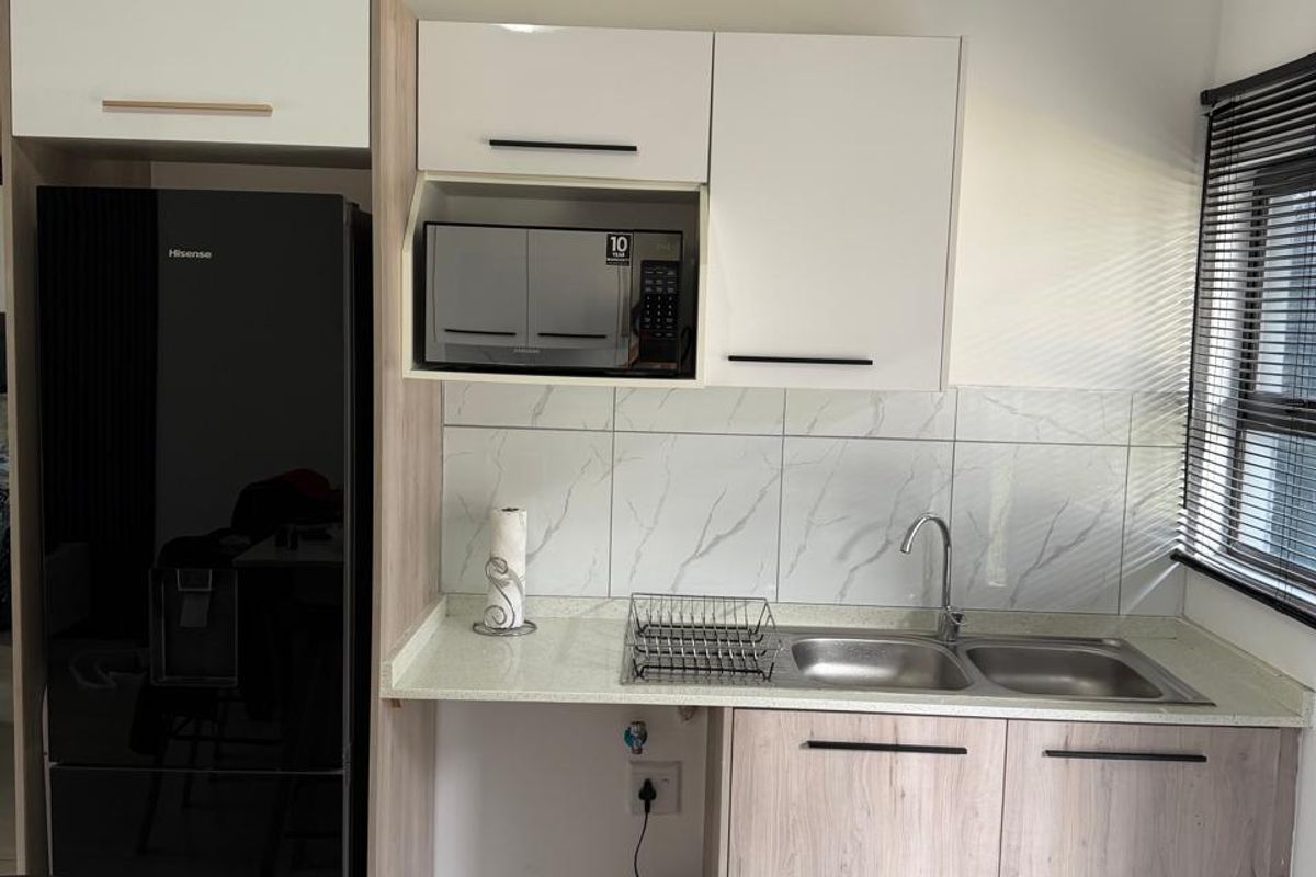 A modern, compact kitchen featuring light wood-grain cabinetry, marble-effect tiled backsplash, double sink, and integrated microwave space.