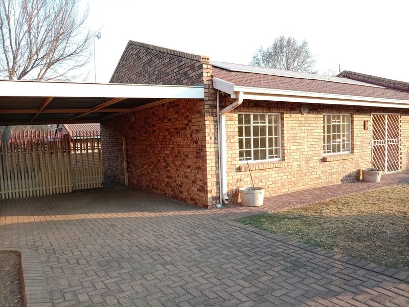 Front elevation of a brick townhouse featuring a paved driveway, attached carport, security gate, and roof-mounted solar panels.