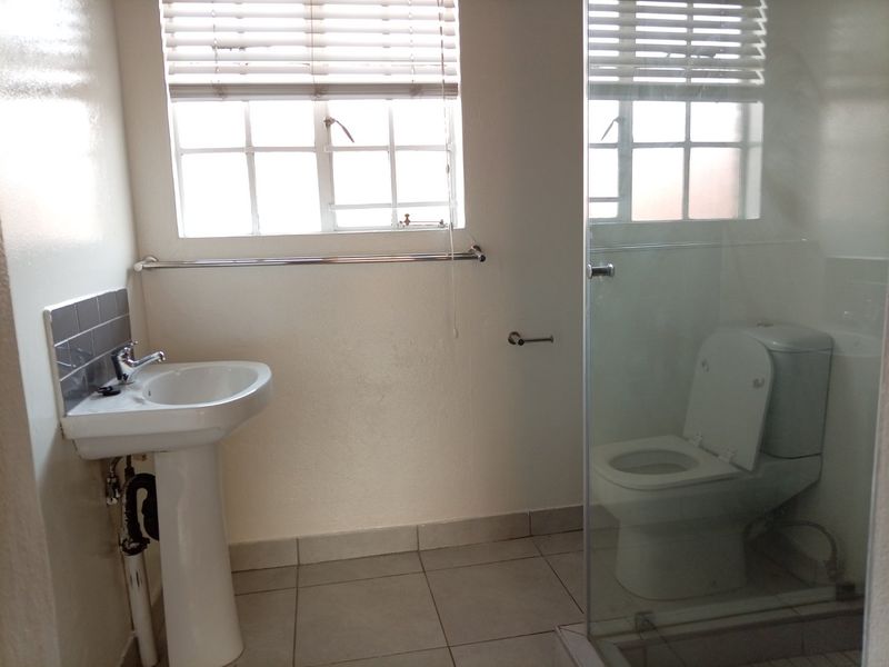 A functional bathroom featuring a pedestal basin, a glass-enclosed shower, and a toilet, finished with neutral floor tiles and a window for natural light.