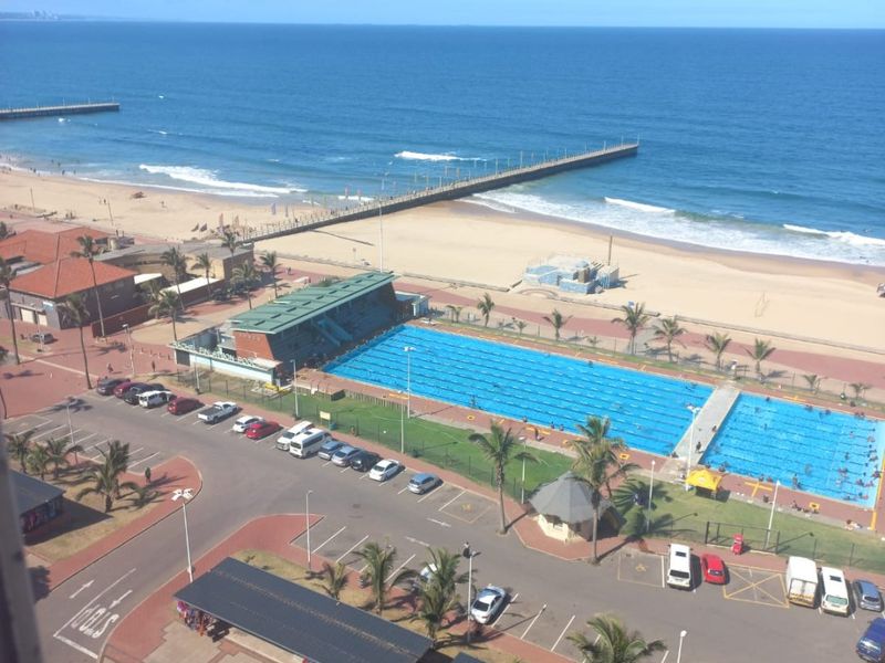 Elevated coastal view overlooking the beach, ocean, and public swimming pool facilities.