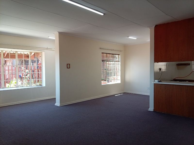 An empty open-plan living and kitchen area featuring dated wooden cabinetry, carpet flooring, and large windows with blinds.
