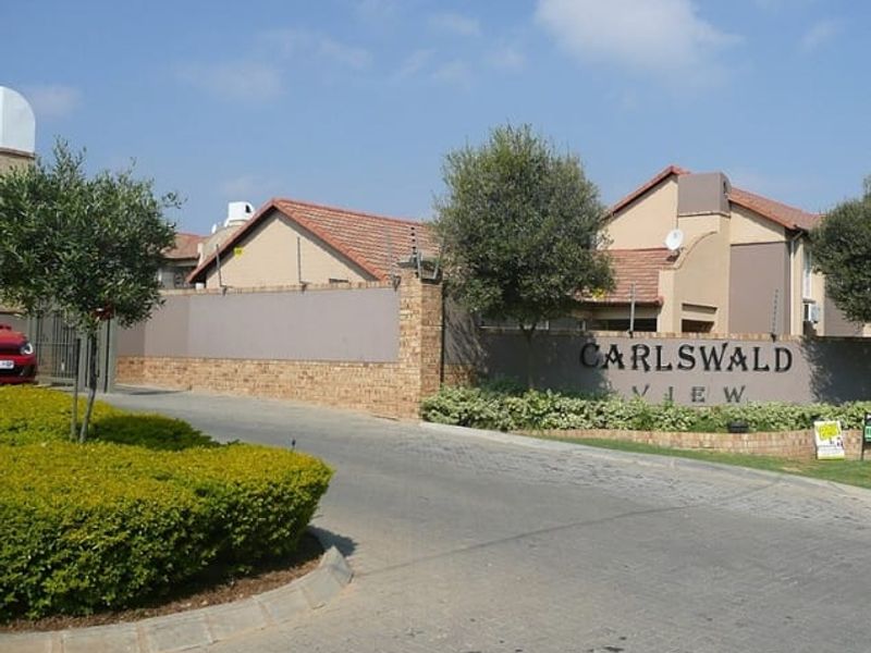 The entrance to Carlswald View apartment complex featuring a paved driveway, brick boundary wall with electric fencing, and established landscaping.