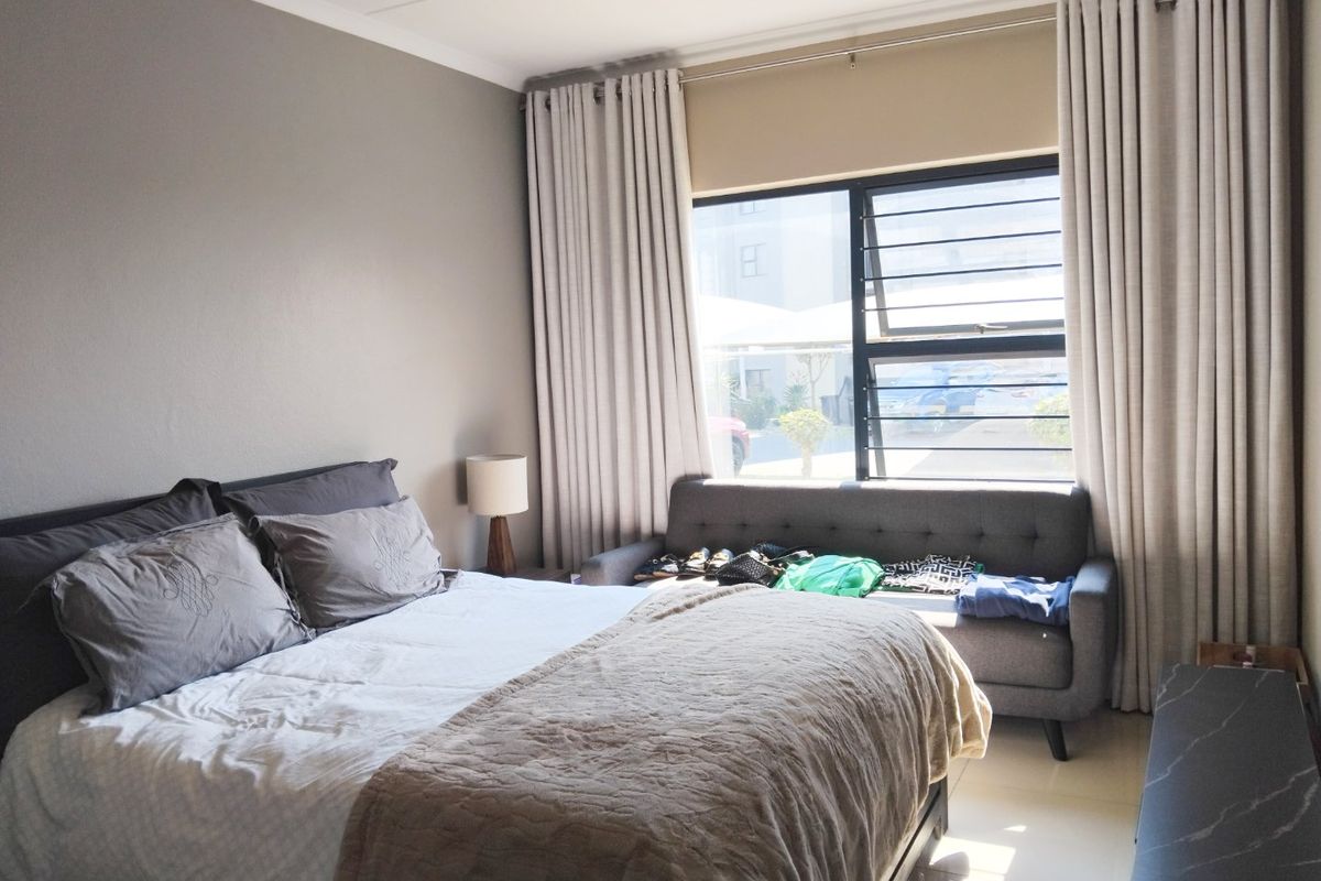 A well-lit bedroom featuring tiled flooring, neutral wall tones, and a large window fitted with security bars and full-length curtains.