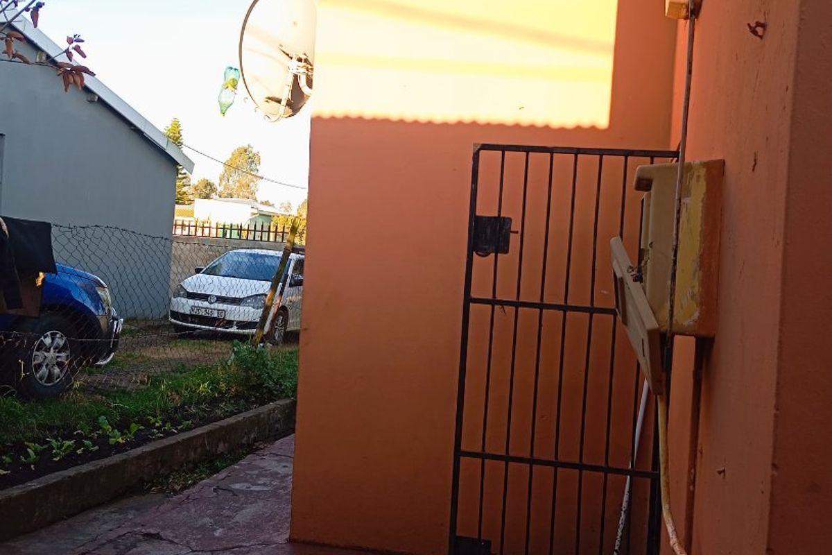 Rear exterior area featuring a metal security gate, concrete paving, and an external electrical meter box against a painted masonry wall.