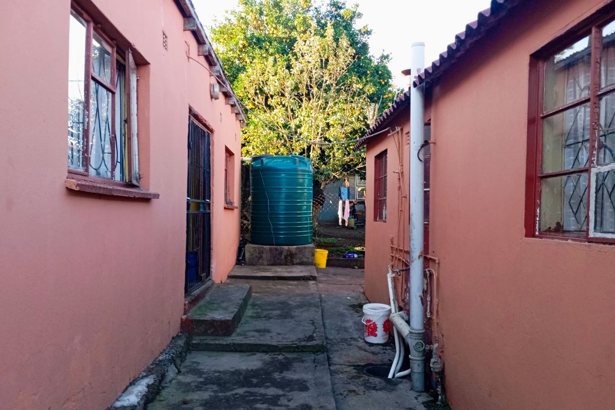 A narrow rear exterior passage between two buildings featuring a water storage tank, concrete paving, and basic utility plumbing.