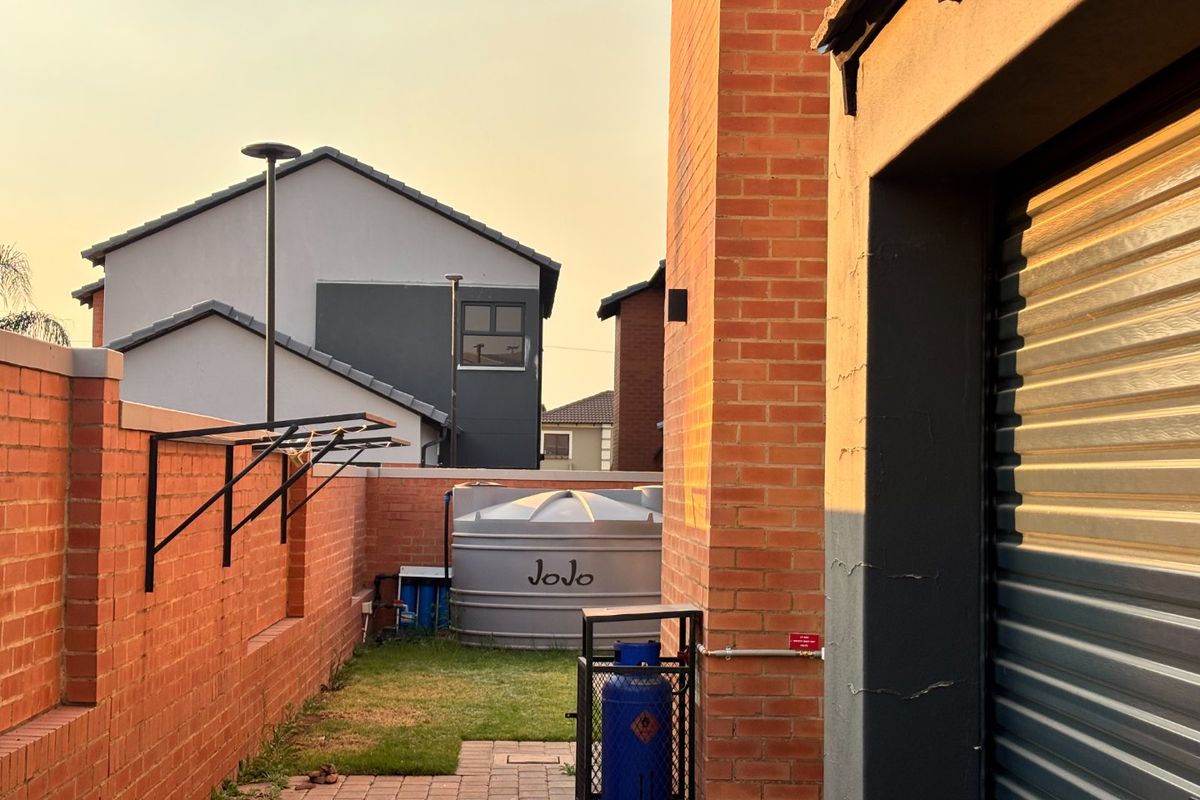 A side exterior view of the townhouse featuring a brick boundary wall, paved path, and a water storage tank adjacent to the garage.