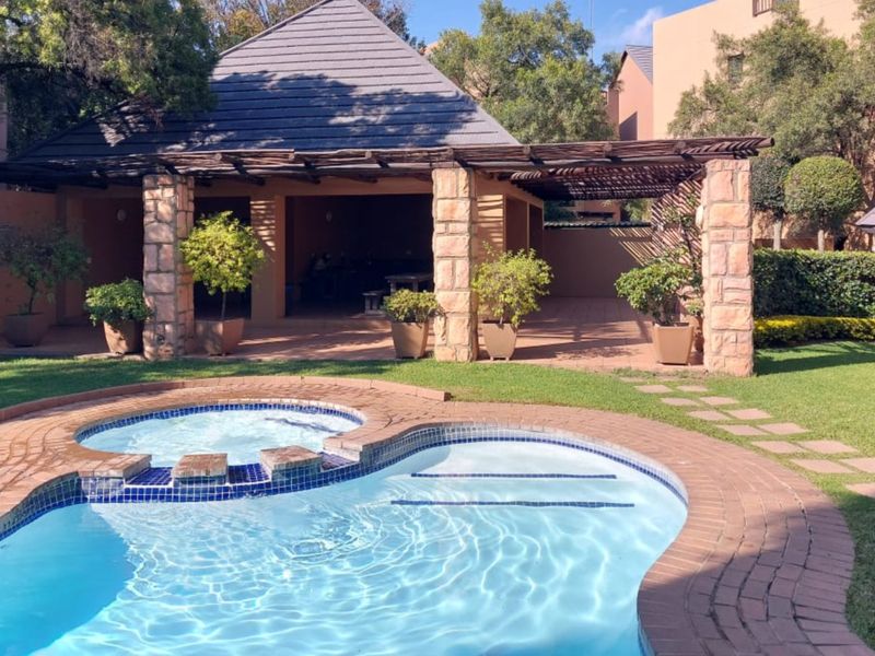 A well-maintained outdoor pool area featuring a swimming pool, integrated spa, and a covered gazebo with stone pillars, set within a landscaped garden.