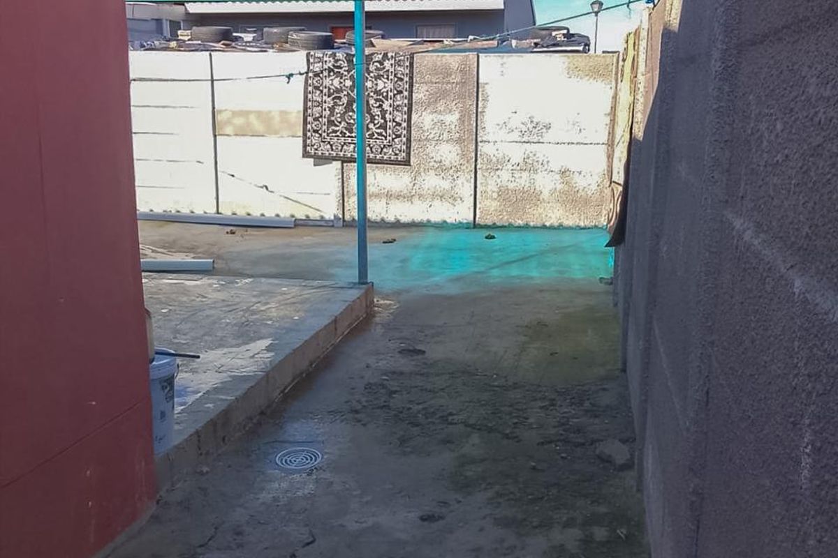 A narrow rear exterior area featuring a paved patio, concrete boundary walls, and a covered section with polycarbonate roofing.