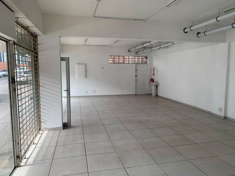 An open-plan commercial space featuring tiled flooring, exposed ceiling services, and a wall-mounted basin, suitable for various business or retail applications.