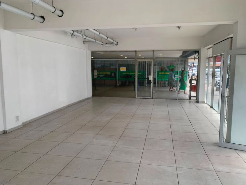 Open-plan commercial retail space featuring tiled flooring, exposed ceiling infrastructure, and a large glass storefront facade with a security gate.