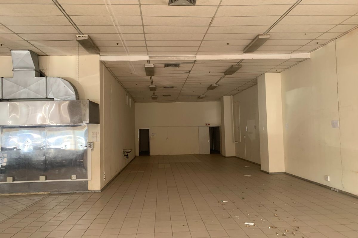 An empty commercial kitchen space featuring a large stainless steel extractor hood, tiled flooring, and a suspended ceiling with integrated lighting.
