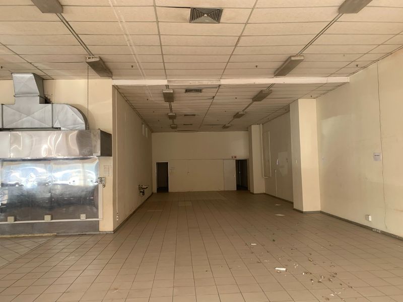 An empty commercial kitchen space featuring a large stainless steel extractor hood, tiled flooring, and a suspended ceiling with integrated lighting.
