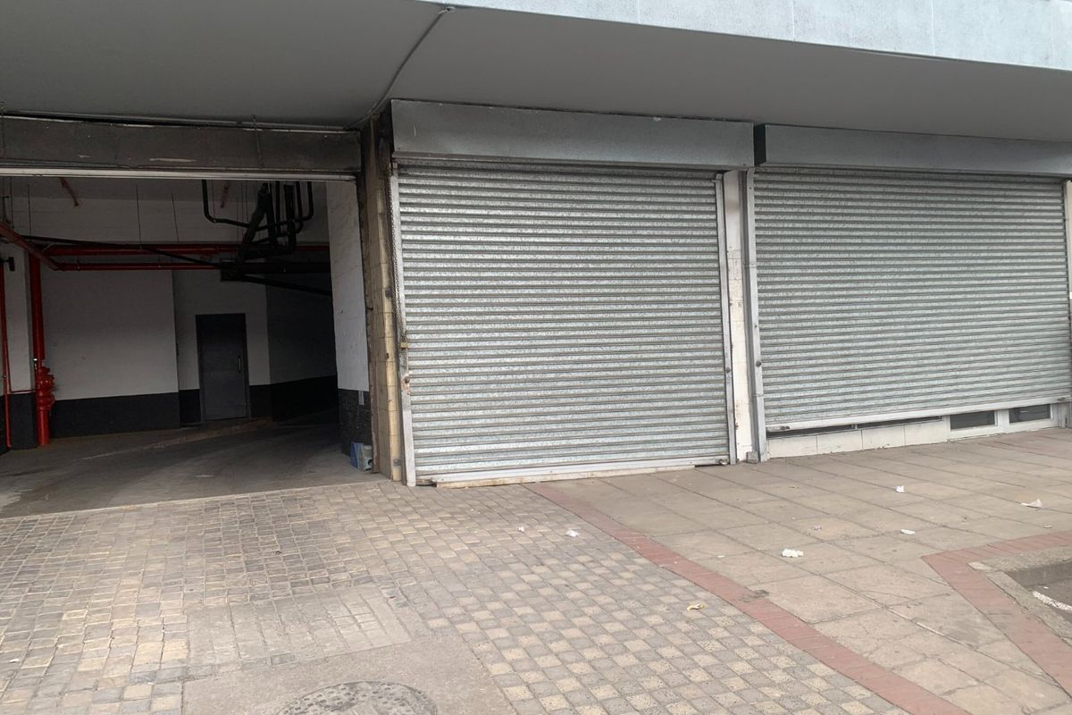 Commercial storefront exterior featuring metal roller shutters and adjacent access to a covered parking or utility area.