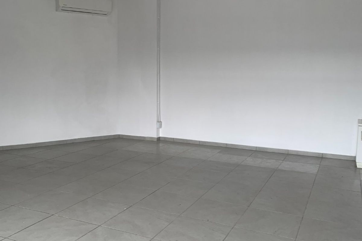 An empty commercial office space featuring ceramic floor tiles, a suspended ceiling with fluorescent lighting, and a wall-mounted split-system air conditioning unit.