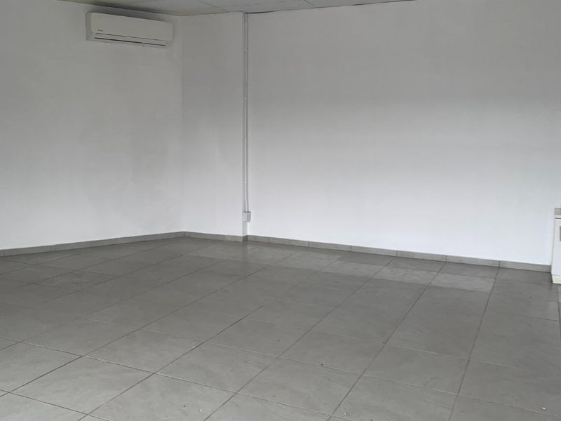 An empty commercial office space featuring ceramic floor tiles, a suspended ceiling with fluorescent lighting, and a wall-mounted split-system air conditioning unit.