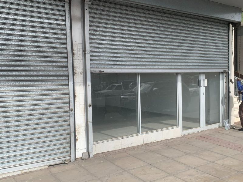 Commercial storefront featuring large glass windows and secure metal roller shutter doors, situated on a paved sidewalk.