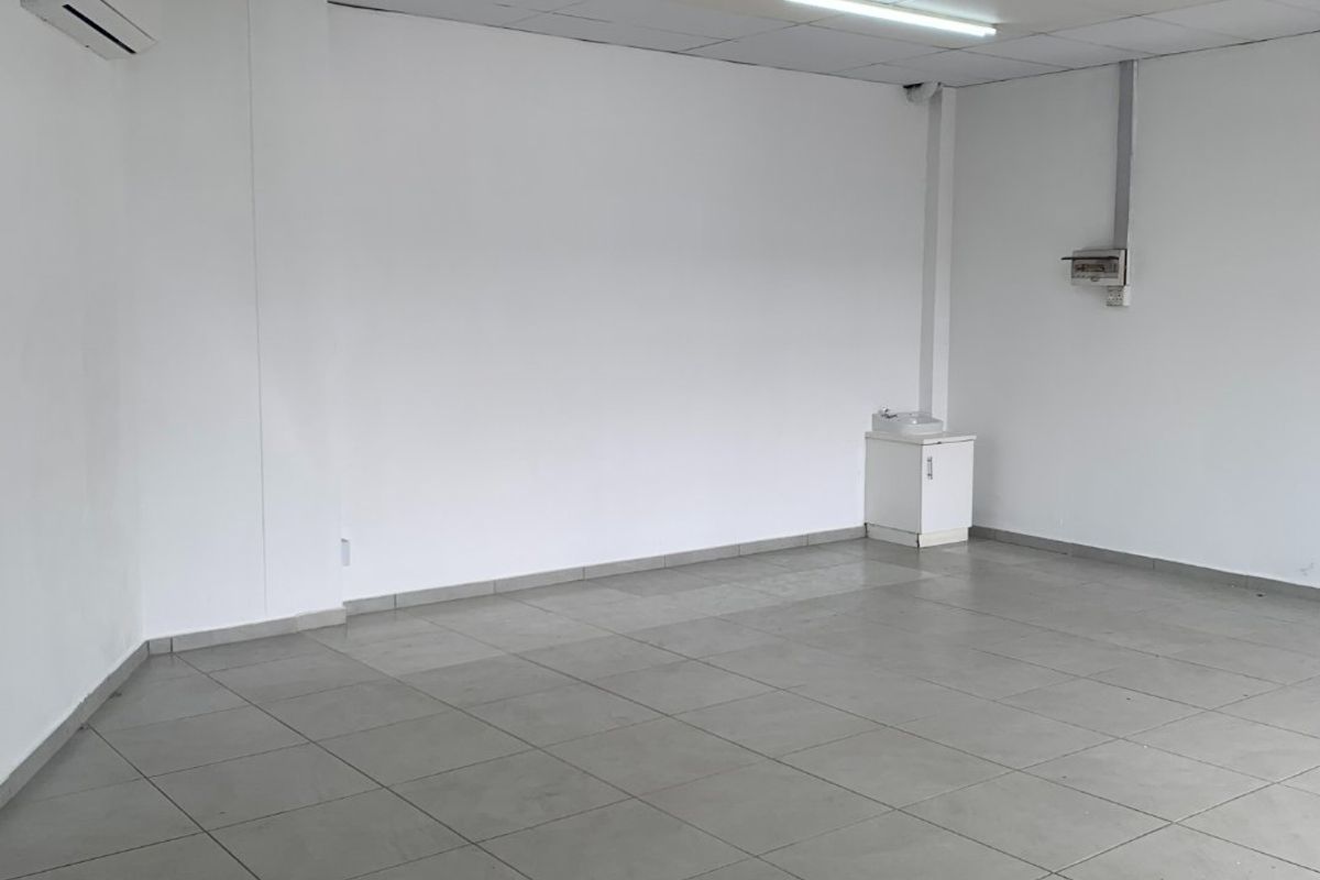 An empty commercial office space featuring tiled flooring, a suspended ceiling with fluorescent lighting, and a wall-mounted air conditioning unit.