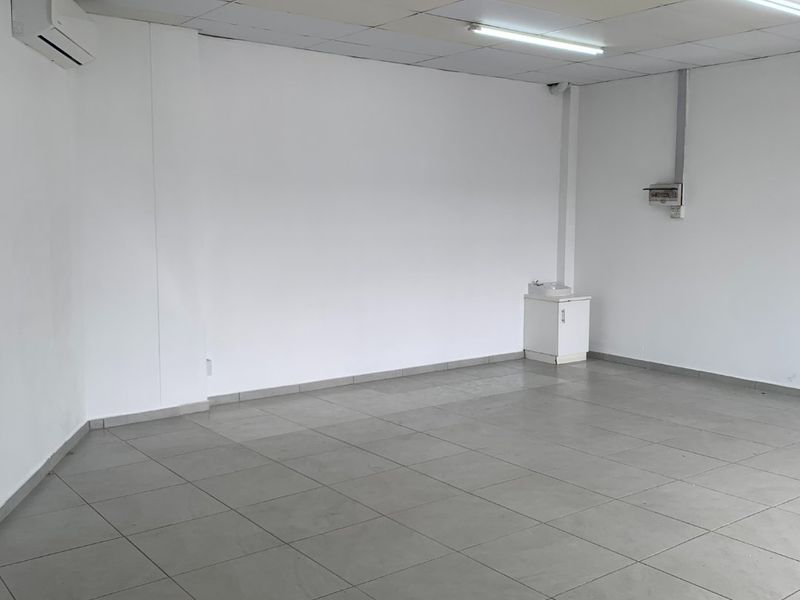 An empty commercial office space featuring tiled flooring, a suspended ceiling with fluorescent lighting, and a wall-mounted air conditioning unit.