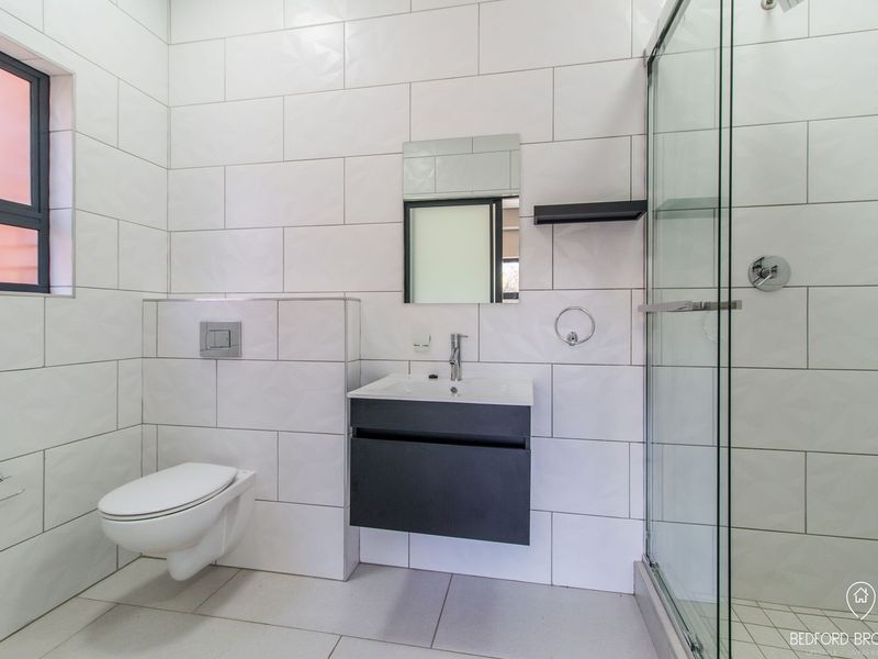 Modern bathroom featuring a wall-mounted toilet, sleek vanity cabinet with basin, and a glass-enclosed shower with contemporary white wall tiling.