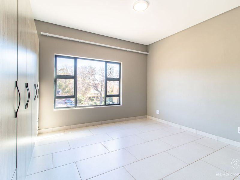 A bright, empty bedroom featuring neutral tiled flooring, built-in cupboards, and a large window providing natural light.