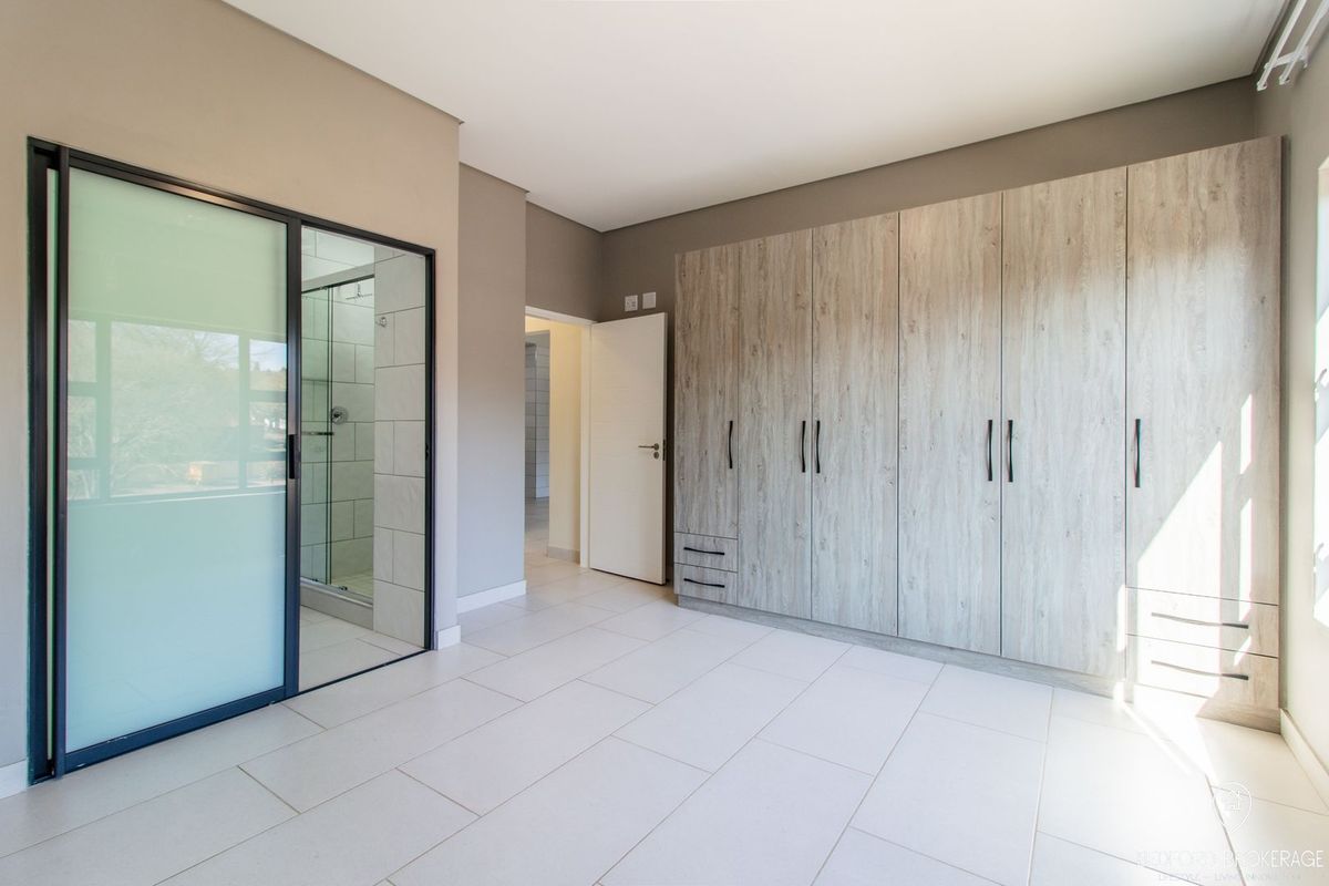 Spacious bedroom featuring modern light wood built-in cupboards, neutral tiled flooring, and a sleek glass-enclosed en-suite bathroom.