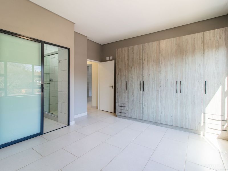 Spacious bedroom featuring modern light wood built-in cupboards, neutral tiled flooring, and a sleek glass-enclosed en-suite bathroom.