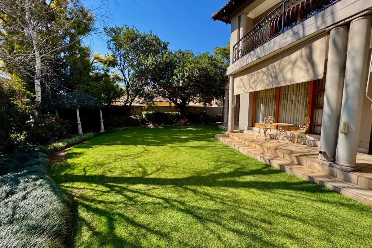 A well-maintained rear garden featuring a manicured lawn, mature trees, and a tiled patio area with a covered balcony above.