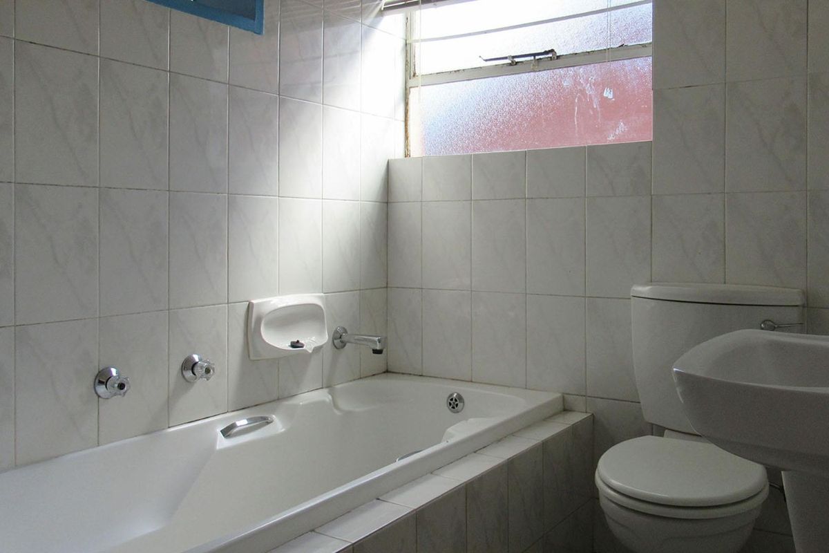 A dated bathroom featuring a built-in bathtub, pedestal basin, and toilet, finished with white ceramic wall tiles and a louvered window.