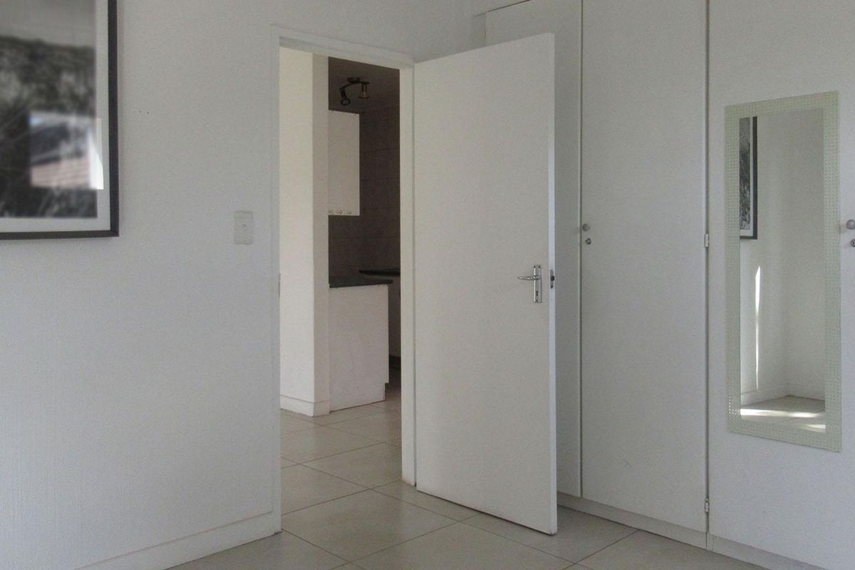 A clean, neutral bedroom featuring built-in cupboards, tiled flooring, and a view through to the adjacent kitchen area.