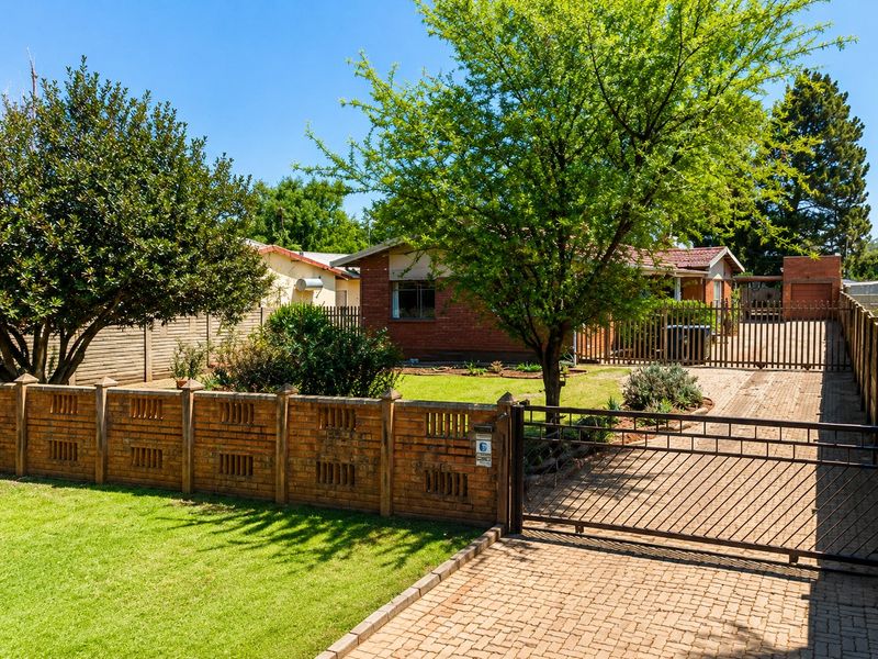 Front view of a brick house featuring a paved driveway, secure metal gate, and established garden with a brick boundary wall.