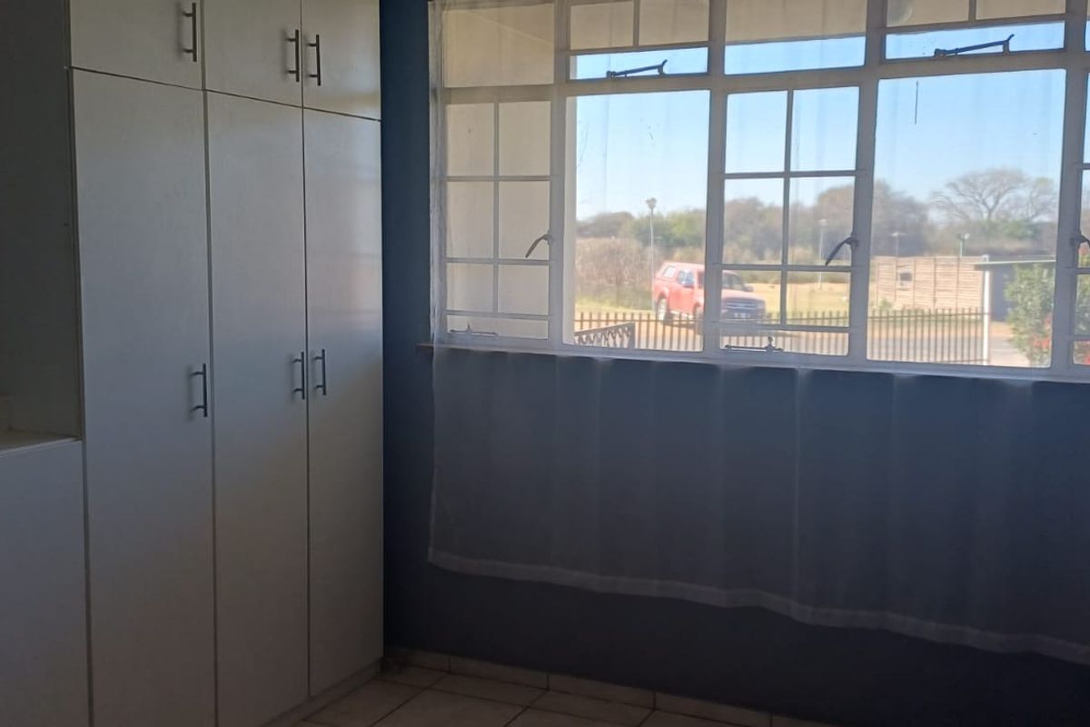 A bright, empty bedroom featuring built-in cupboards, tiled flooring, and a large window providing natural light and a view of the exterior.