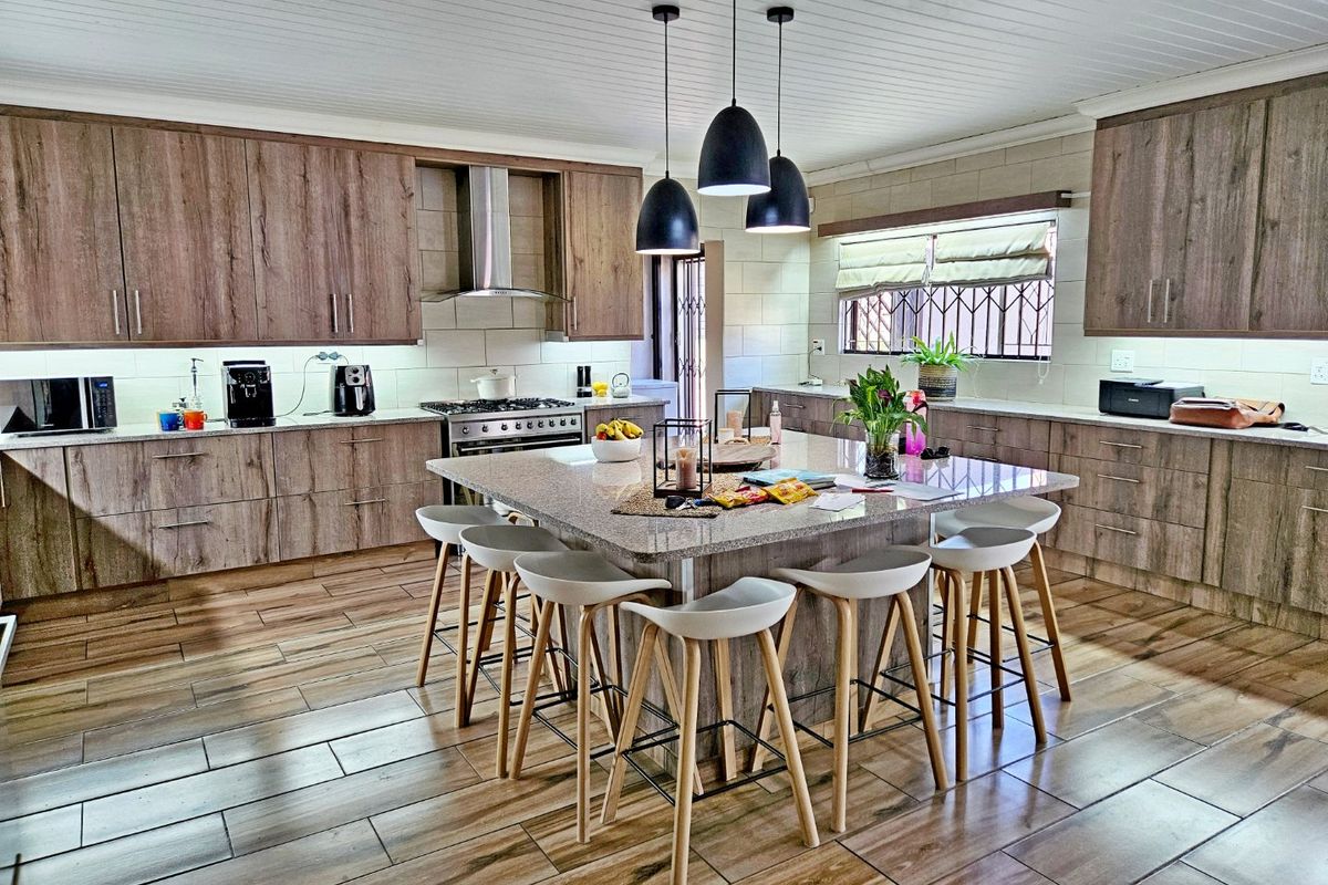 Spacious modern kitchen featuring a large central island with granite countertops, ample wood-finish cabinetry, stainless steel appliances, and contemporary pendant lighting.