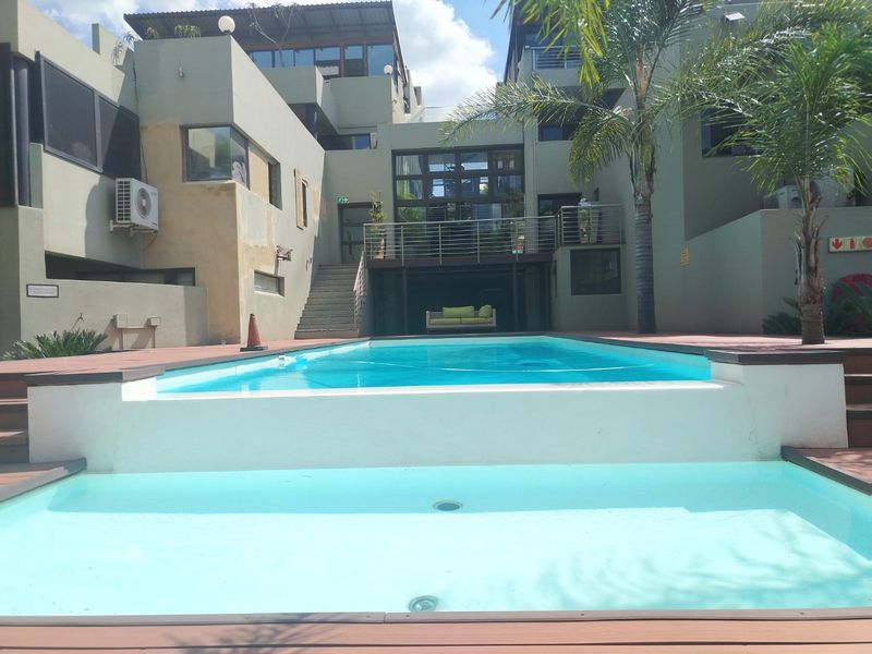 A communal pool area featuring timber decking, a raised pool structure, and access stairs leading to the townhouse complex buildings.