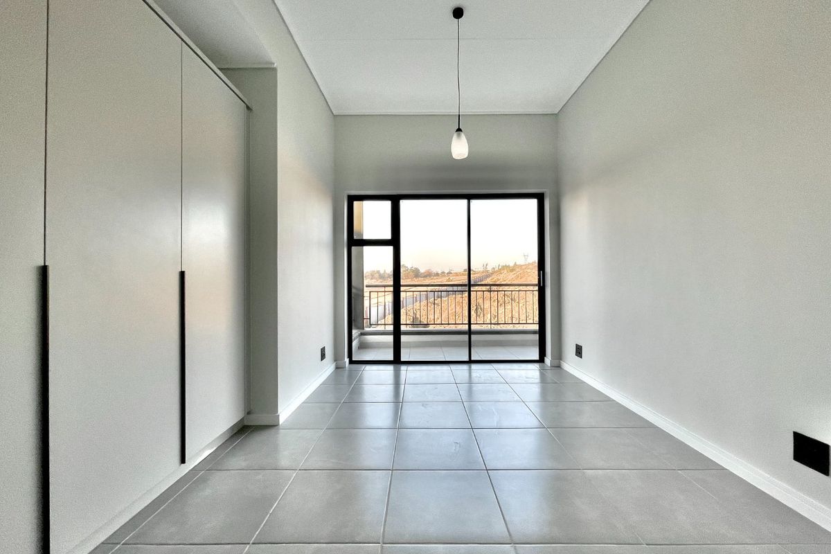 A bright, modern bedroom featuring neutral tiled flooring, ample built-in cupboard space, and a sliding glass door leading to a private balcony.